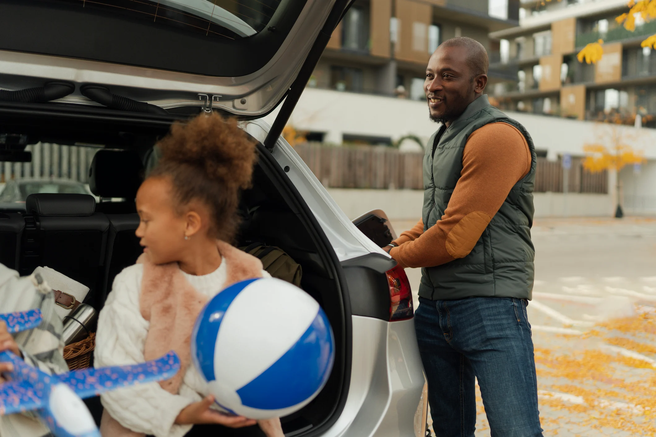 A person loads items into the open boot of a car parked on a residential street, with another person holding a ball nearby on an autumn day.