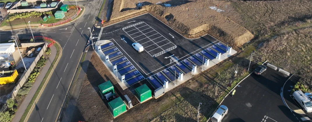Aerial view of a  large-scale EV charging site with multiple charging bays, green utility units, and surrounding infrastructure under development.