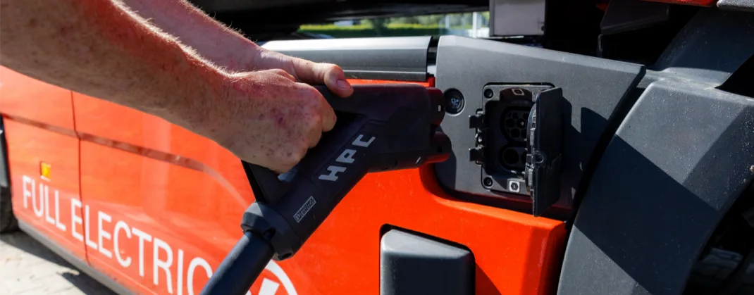 A close-up of hands plugging a high-power charging (HPC) connector into the charging port of a red fully electric vehicle.