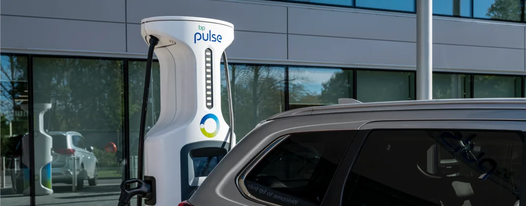 A bp pulse electric vehicle charger connected to a gray car, set against a modern office building with reflective glass windows.