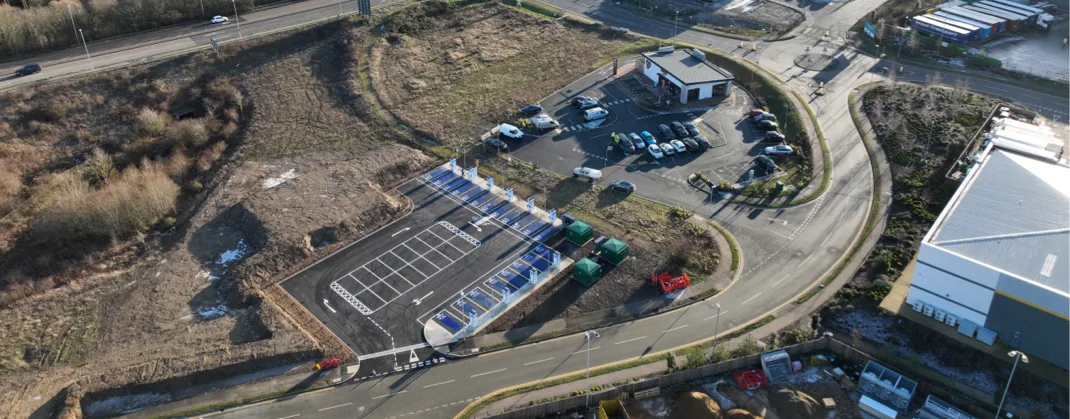An aerial view of newly constructed electric vehicle charging infrastructure, with multiple charging bays, a parking area, and a nearby building surrounded by open land and roads.