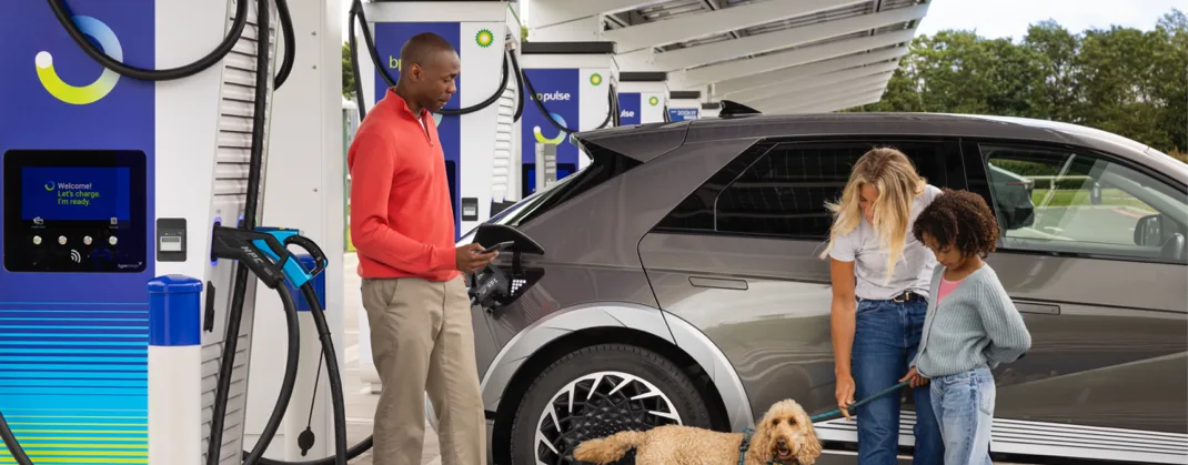 An EV driver plugs in their vehicle at a bp pulse charging station while a family with a dog waits nearby, relaxing as their car charges.