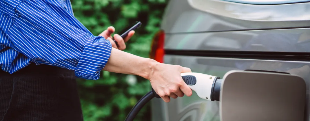 A person charging their EV vehicle with a charger in one hand and a mobile phone in another.