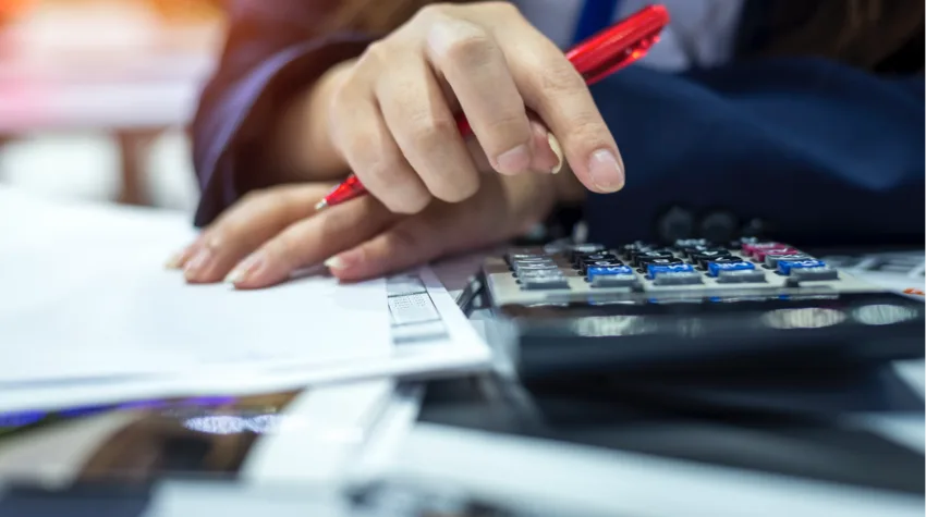 A woman's hand holding a red pen, and hovering above a calculator that she is about to use. 