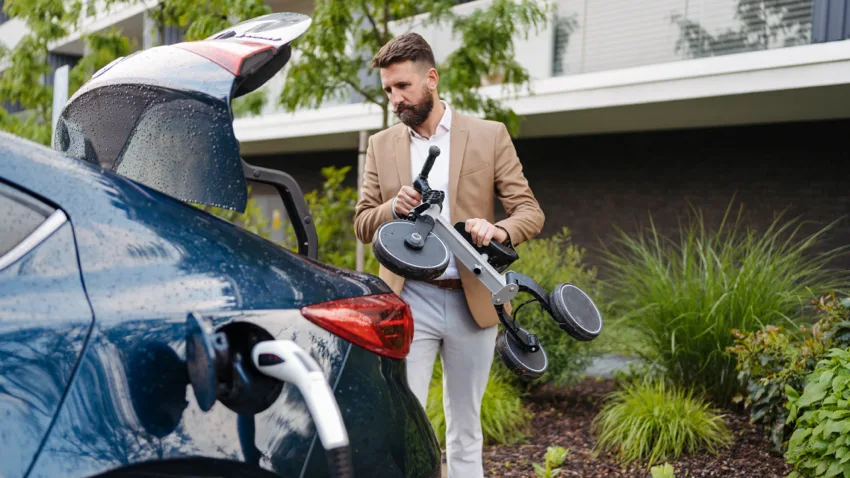 The rear of a dark blue Electric Vehicle is visible,  a man is carrying a tricycle and is putting it into the open boot. It is a rainy day and water droplets can be seen on the car.
