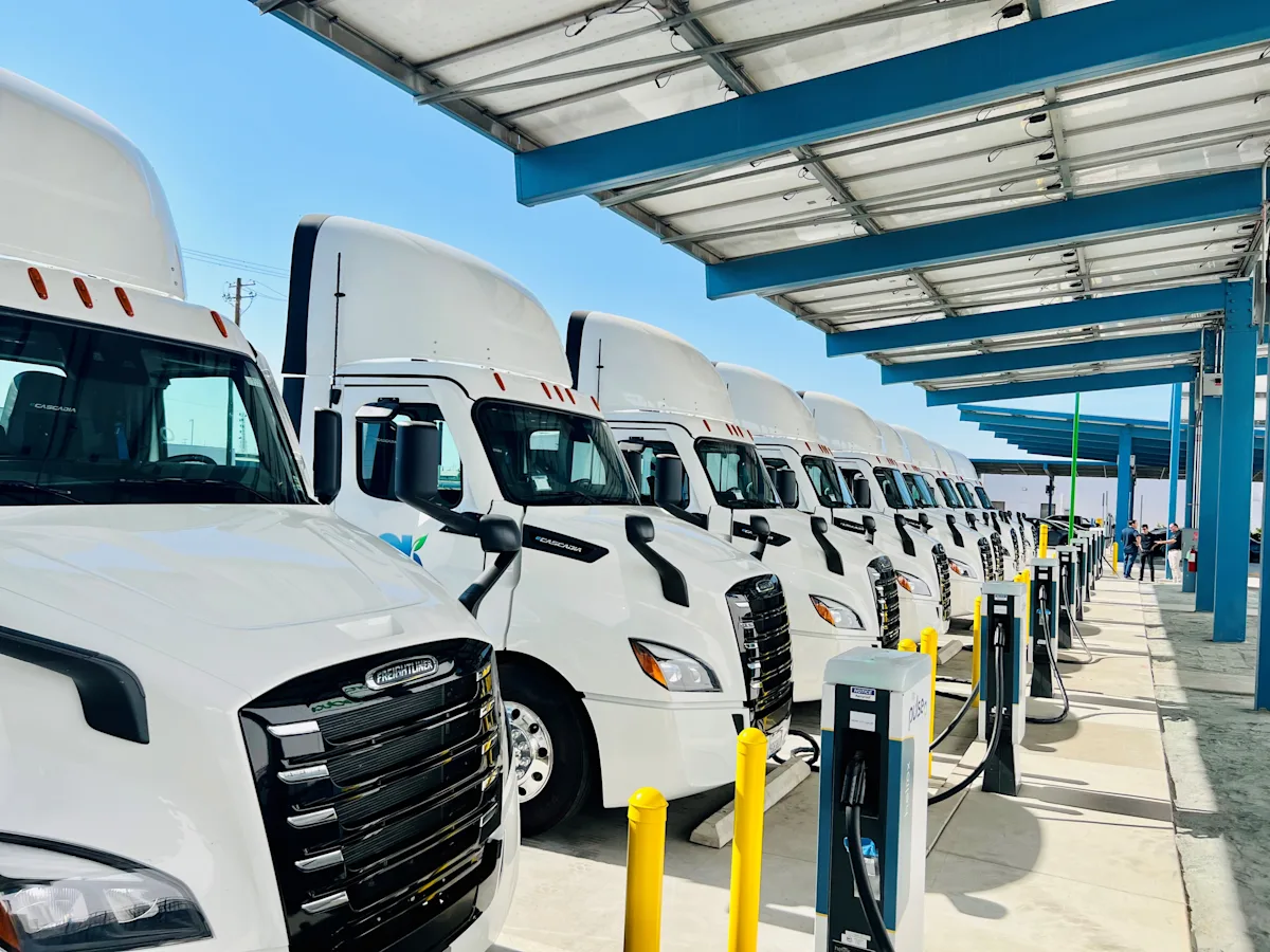 A row of white electric trucks are charging on bp pulse chargers on a bright day. 