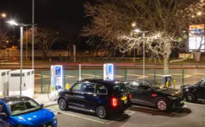A charging station with four charging bays all occupied with Electric Vehicles. There is a dark sky, illuminated with street lighting. A bare tree and signage are visible.