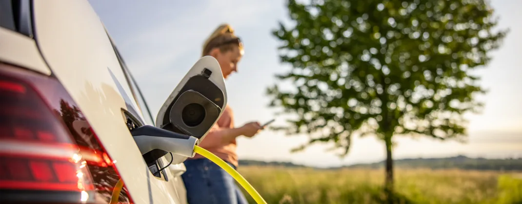 A woman browsing on her phone right next to her EV in an open green field on a sunny day, while the EV is being charged. 