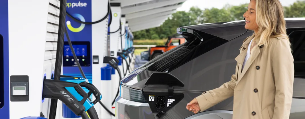 A woman in a tan jacket is smiling as she walks to the bp pulse charging point to start charging her ev. 