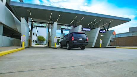 A parking area at an airport terminal with clear directional signage and marked parking spaces. The scene includes a multi-level parking structure in the background and a well-maintained roadway leading to the terminal. The sky is partly cloudy, adding a dynamic element to the setting.