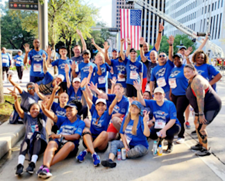 A large group of people in matching blue shirts pose at an 5k outdoor event, some sitting and some standing, raising their hands and smiling