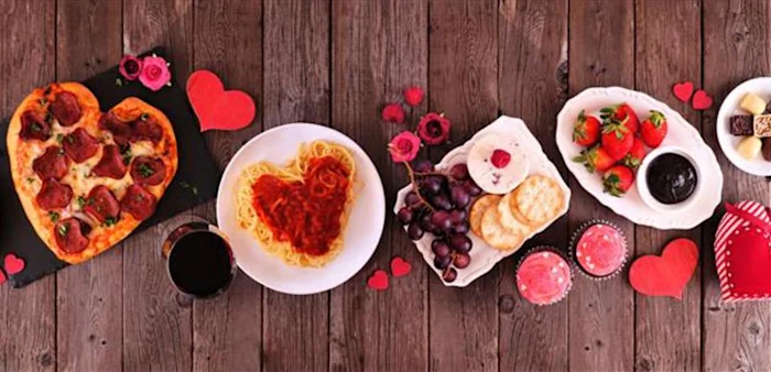 Overhead view table setting on a dark wood banner background. Heart shaped pizza, pasta, wine, cheese plate and desserts.