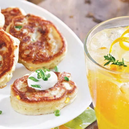 A plate of mini-pancakes with bacon, cheddar goat cheese and corn with a glass of juice