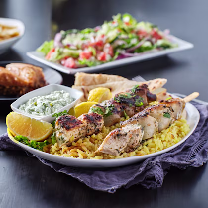 Two chicken souvlaki skewers in a plate sitting on top of yellow rice with a side of Tzatziki sauce and lemon wedges