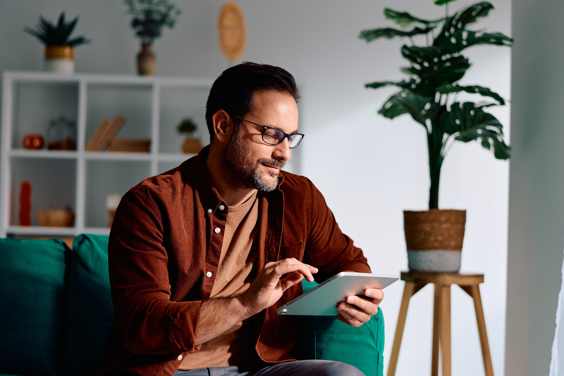 Un homme avec des lunettes est sur son canapé vert, il consulte sa tablette pour identifier la banque qui proposent des frais bancaires les moins élevés du marché.