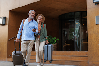 Couple de voyageurs seniors souriants quittant sereinement leur hôtel avec des valises, illustrant la sérénité apportée par l’assurance voyage incluse avec leurs cartes bancaires.