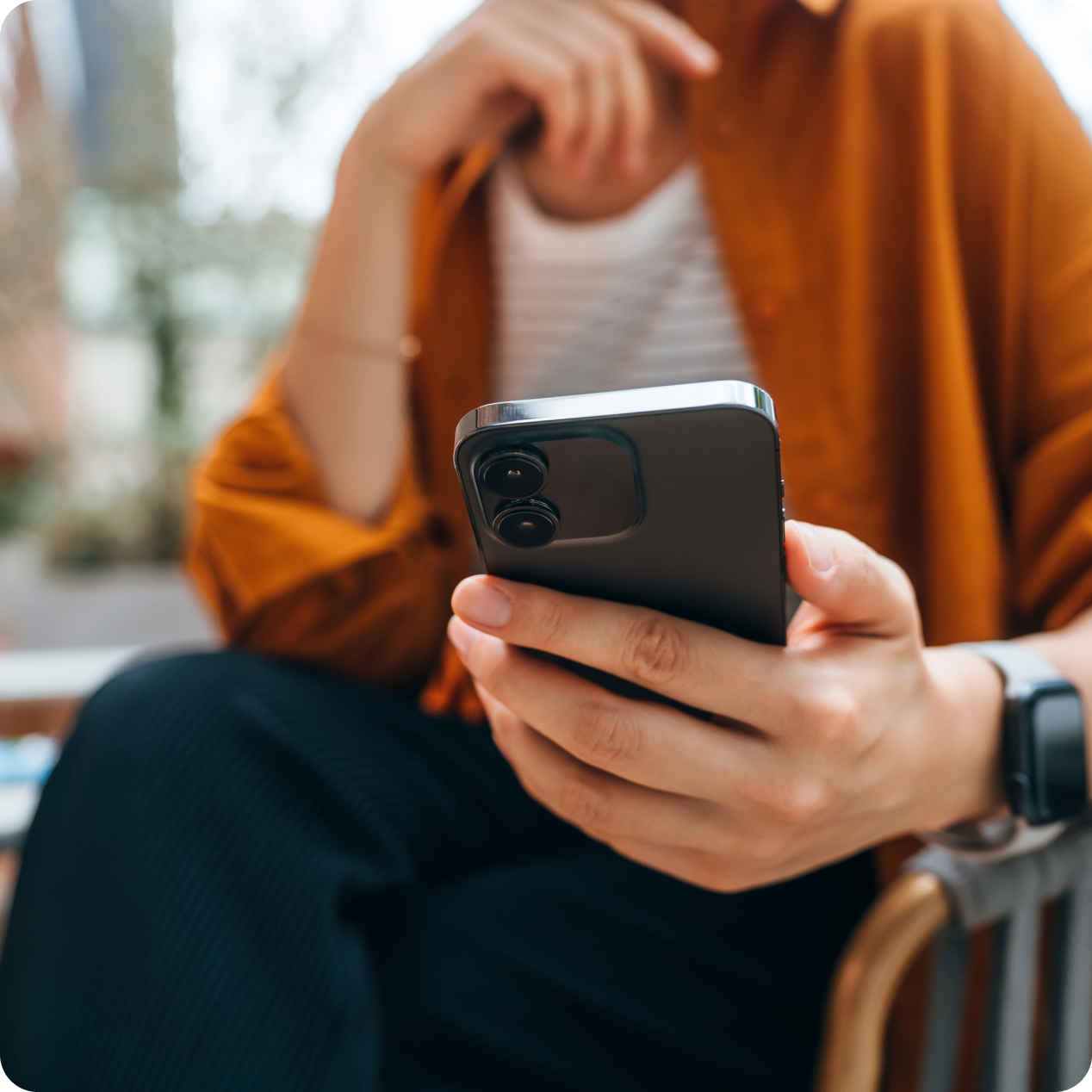 Personne assise tenant un smartphone noir, portant une chemise orange et une montre connectée, en extérieur.