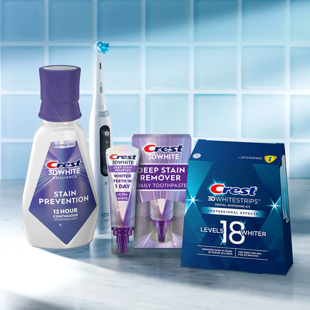 Crest 3D White oral care display: stain-prevention mouthwash, deep-stain remover toothpaste, whitening strips box and an electric toothbrush on a bathroom counter.