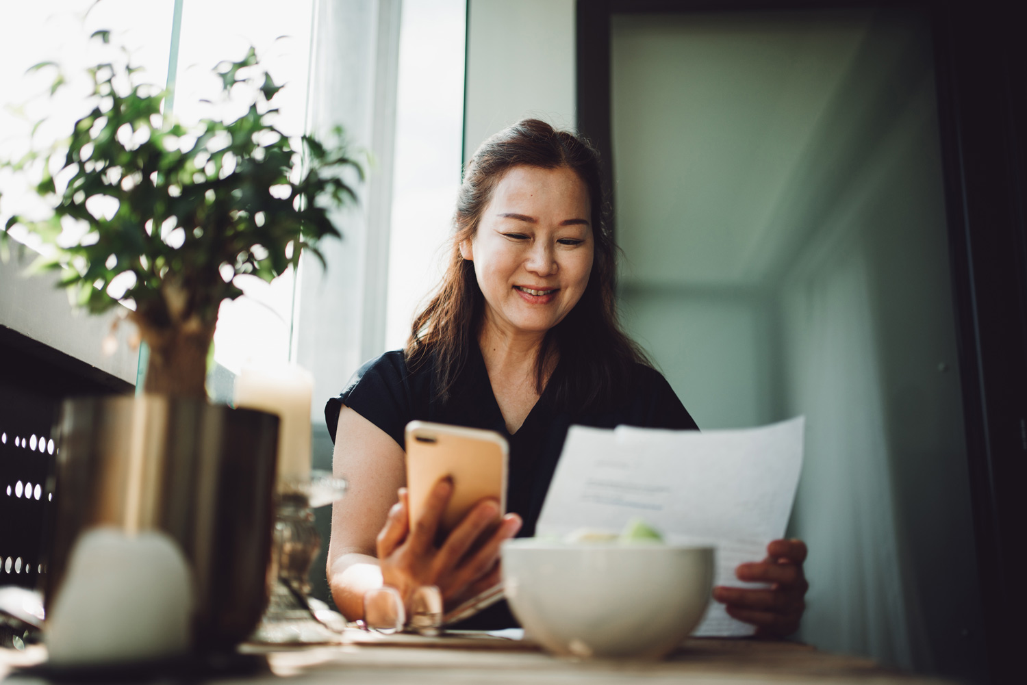 A middle-aged Asian woman looking at her phone and a bank statement while sitting at her kitchen table.