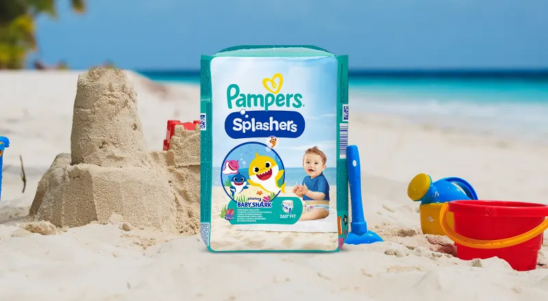 Un paquet de Pampers Splashers Baby-Shark sur une plage avec un château de sable et des jouets de plage