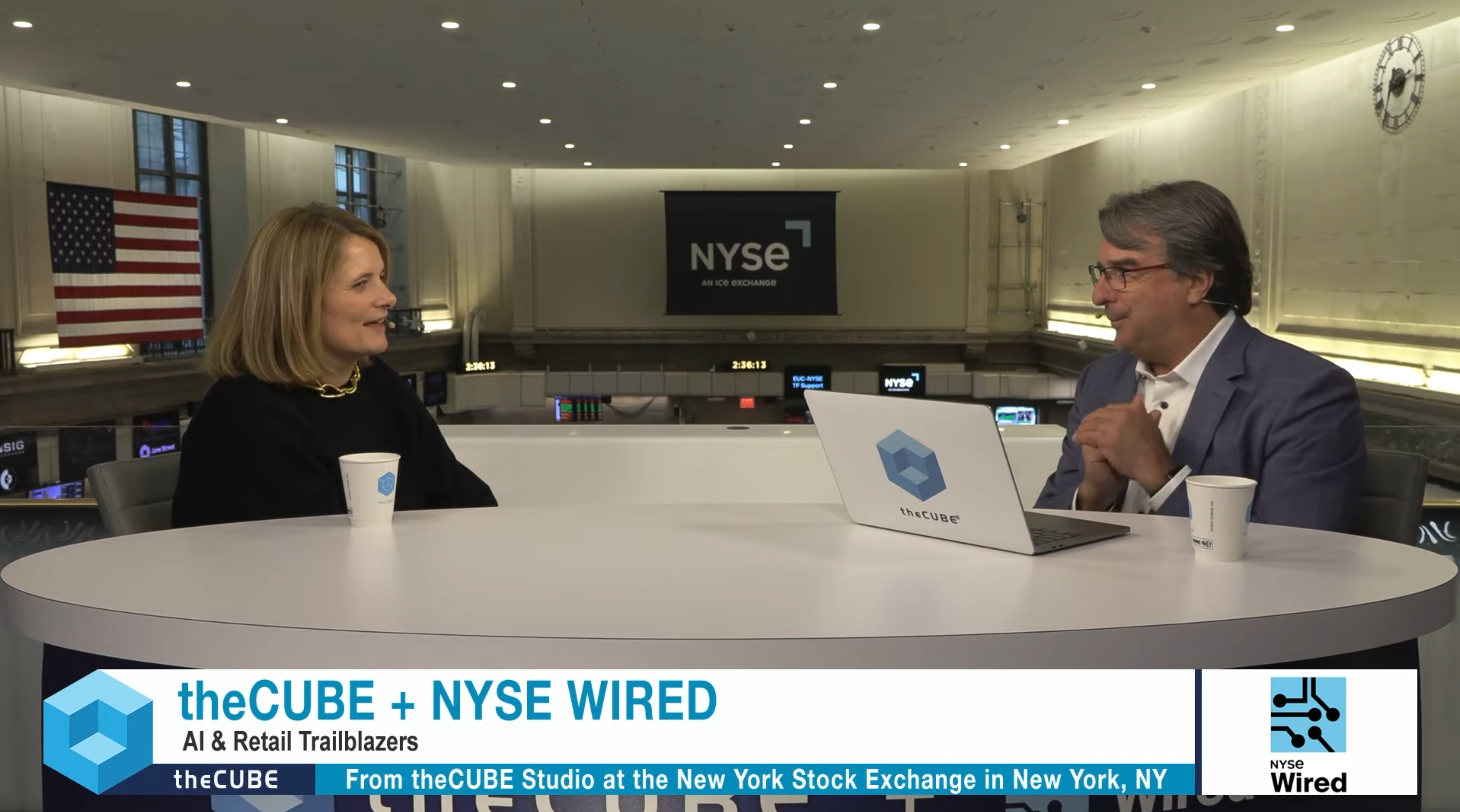 Mirakl Chief Data and AI Officer Anne-Claire Baschet (left) and theCUBE host John Furrier (right) sit at a white table during an interview at the NYSE studio. The background features the New York Stock Exchange trading floor balcony, an American flag, and a screen displaying the NYSE logo. A graphic at the bottom reads "theCUBE + NYSE WIRED | AI & Retail Trailblazers".