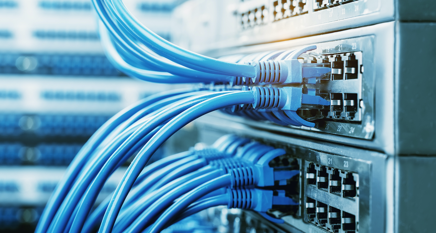 A close-up view of numerous blue Ethernet cables plugged into network switches, with rows of blinking lights visible on the server rack in the background.