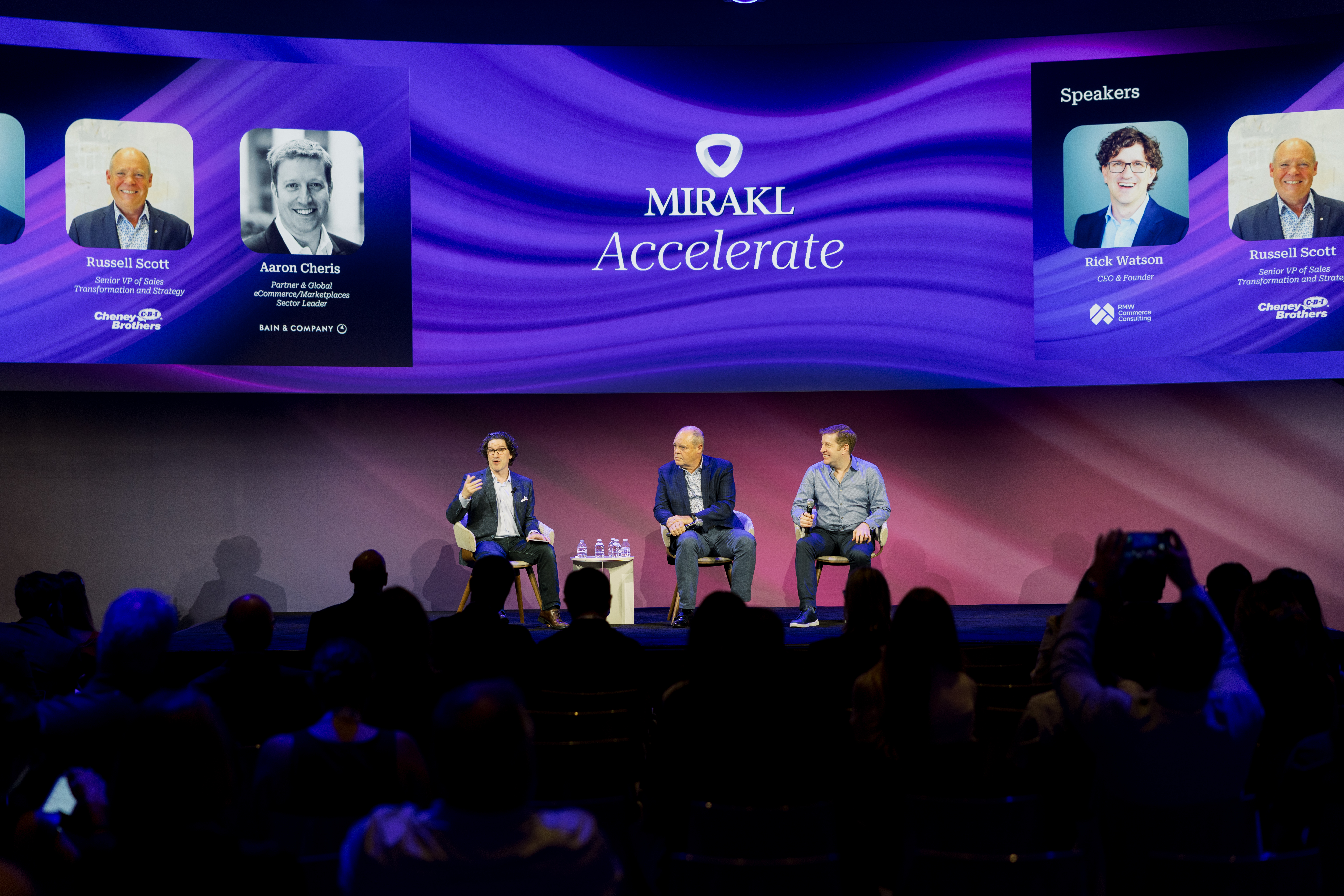 A wide shot of a stage at a conference, featuring three male speakers seated in chairs. The man on the left, wearing a dark jacket, is gesturing with his hands while speaking. The man in the center is wearing a blue jacket, and the man on the right is in a grey long-sleeved shirt. Behind them, a large screen displays the Mirakl Accelerate logo in the center. On either side of the screen, there are headshots and names of "Russell Scott" and "Aaron Cheris" on the left, and "Rick Watson" and "Russell Scott" on the right. The audience is visible in the foreground, with several people holding up phones to take pictures.