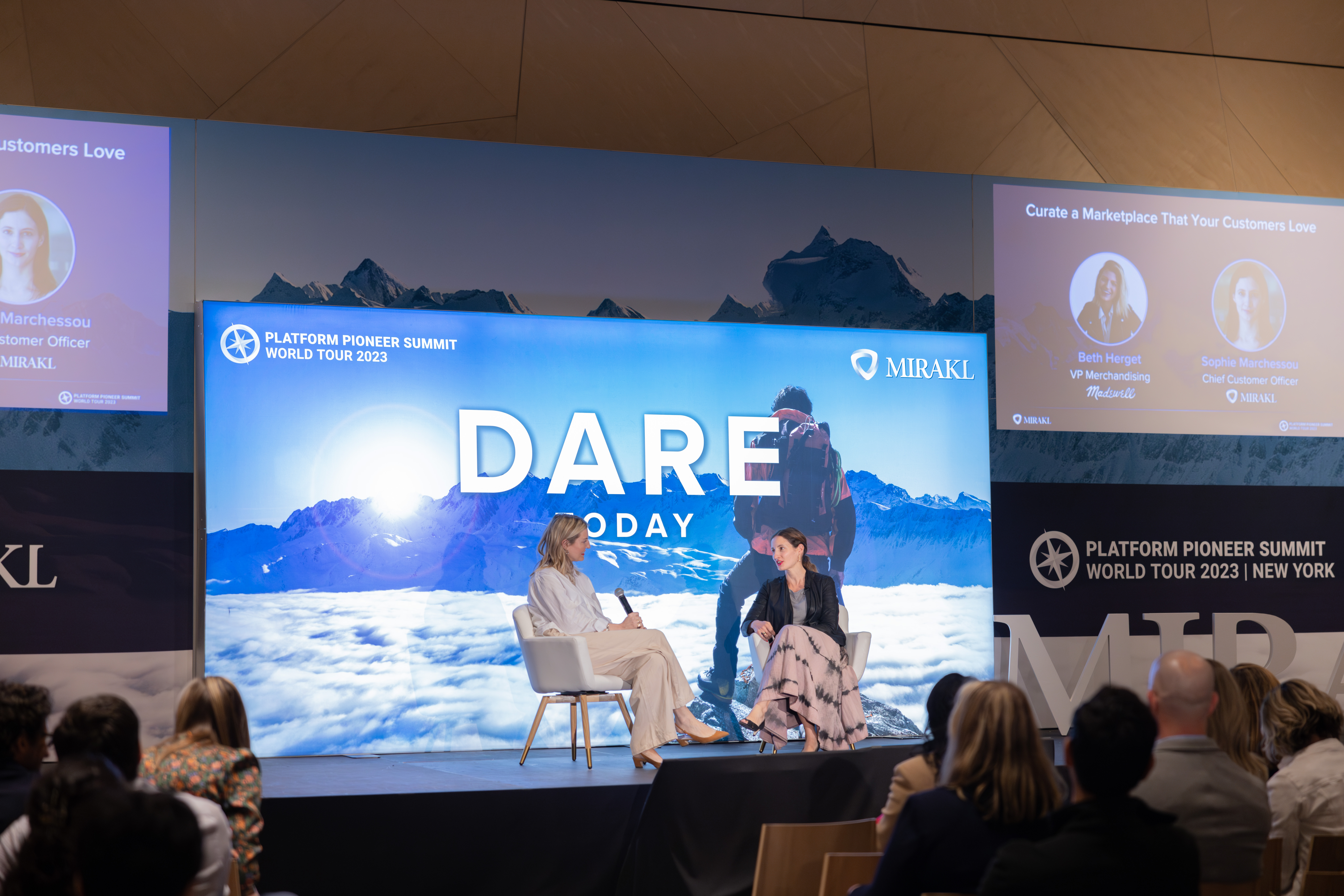 Two women are seated on a stage, engaged in a discussion at a conference. The large central screen behind them displays "DARE TODAY" over a mountain graphic, along with logos for "PLATFORM PIONEER SUMMIT WORLD TOUR 2023" and "MIRAKL". Smaller side screens show speaker profiles. The backs of the audience members are visible in the foreground.