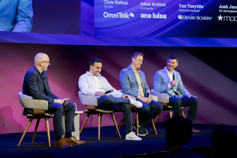 A panel of four men sits on a stage. From left to right, a man with glasses and a beard in a dark suit, a man in a striped white shirt and jeans holding a microphone, a man in a textured blue jacket, and a man in a light blue suit holding a microphone. All are smiling and engaged in conversation. The background screen displays their names and titles, including Chris Walton, Adam Bohbot, Yan Tseytlin, and Josh Janos. Company logos for OmniTalk, Ana Luisa, Henry Schein, and Macy's are also visible.