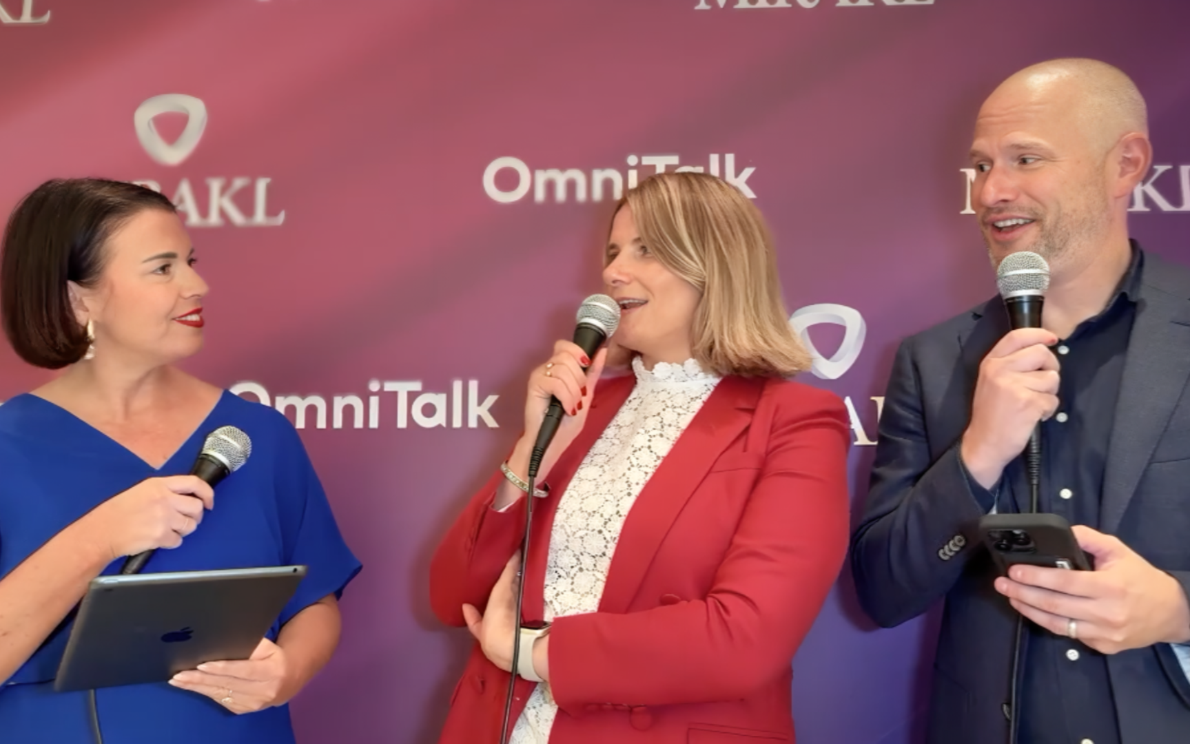 Three individuals are standing against a vibrant purple backdrop adorned with "MIRAKL" and "OmniTalk" logos. A woman on the left, wearing a royal blue top, holds a microphone and a tablet. In the center, another woman in a red blazer over a white lace top also holds a microphone, looking towards the right. On the far right, a bald man in a dark suit holds a microphone in one hand and a smartphone in the other, smiling and looking to his left. All three appear to be engaged in a live discussion or interview.