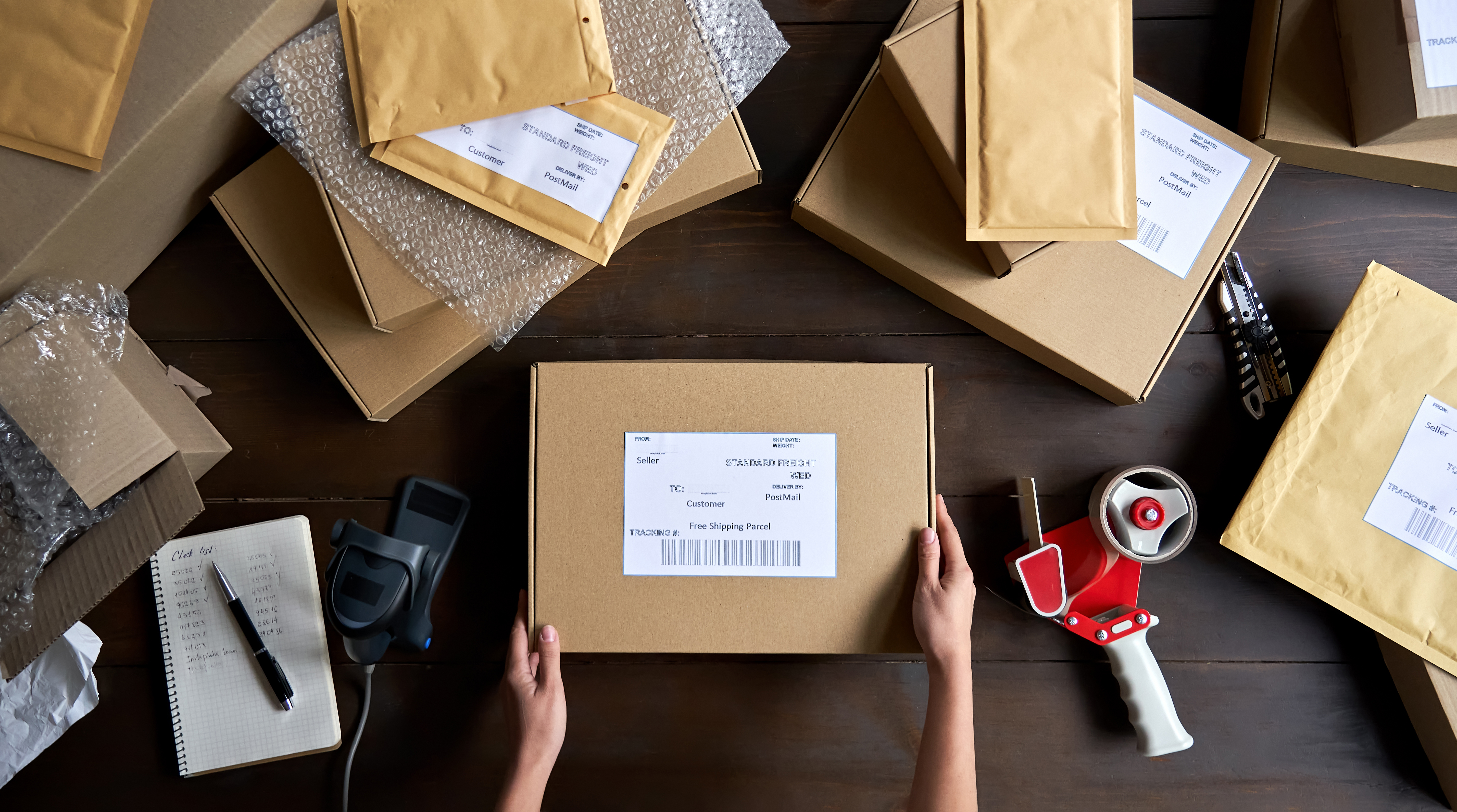 An overhead, flat-lay view of a person's hands holding a cardboard box with a shipping label, ready for dispatch. The wooden table underneath is cluttered with various packing materials, including other boxes, padded envelopes, a tape dispenser, and a barcode scanner.