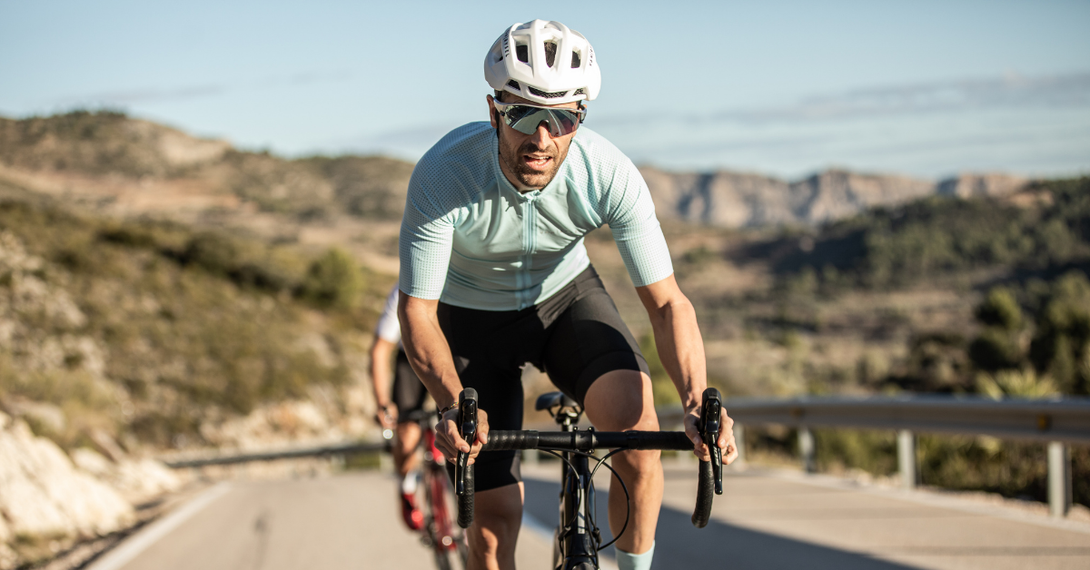 A male cyclist in a light blue jersey, white helmet, and sunglasses leans forward on his road bike, riding intensely on a paved road. The background features a dry, hilly, and mountainous landscape under a clear sky. Another cyclist is partially visible behind him.