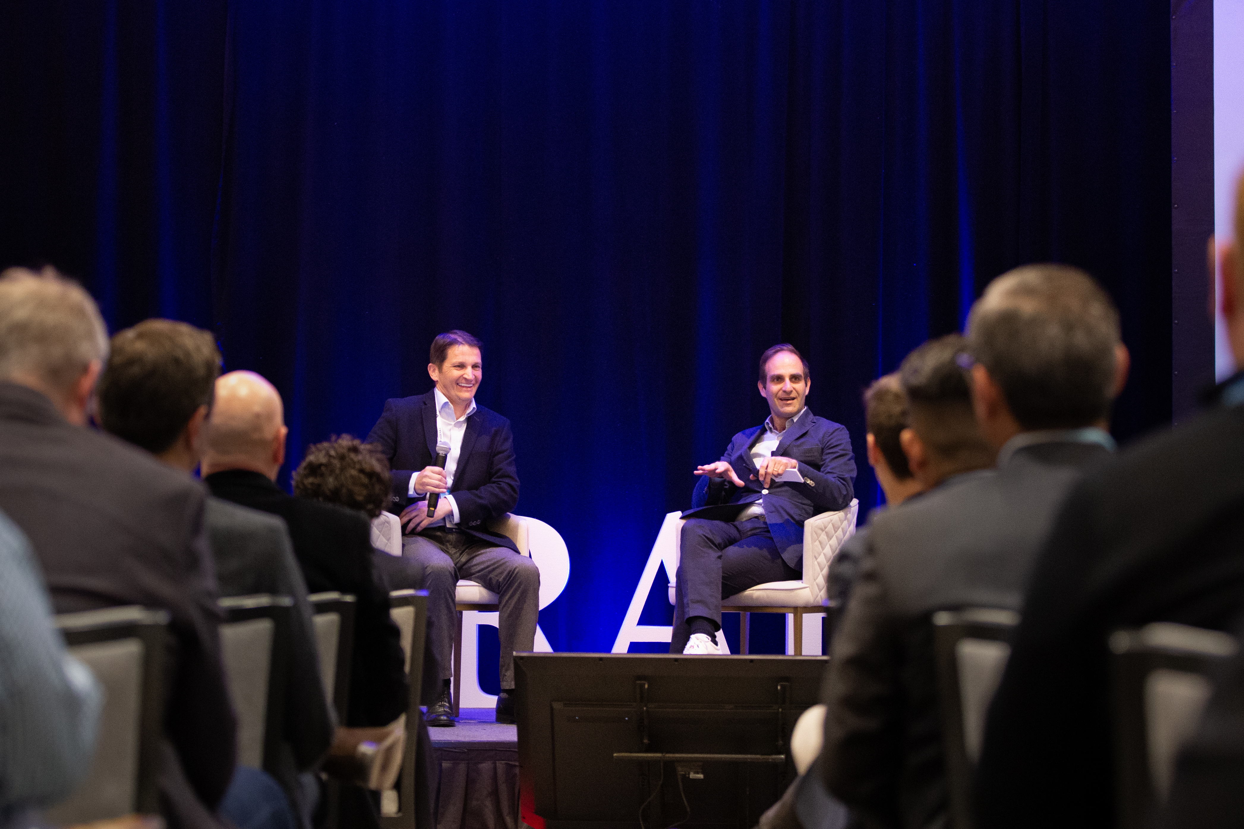Two men are seated on a stage, engaging in a discussion in front of a dark blue curtain backdrop. Both are dressed in business casual attire. In the foreground, the backs of audience members' heads are visible, indicating they are watching the presentation. Large white letters spelling out "RA" are partially visible on the stage floor in front of the speakers.