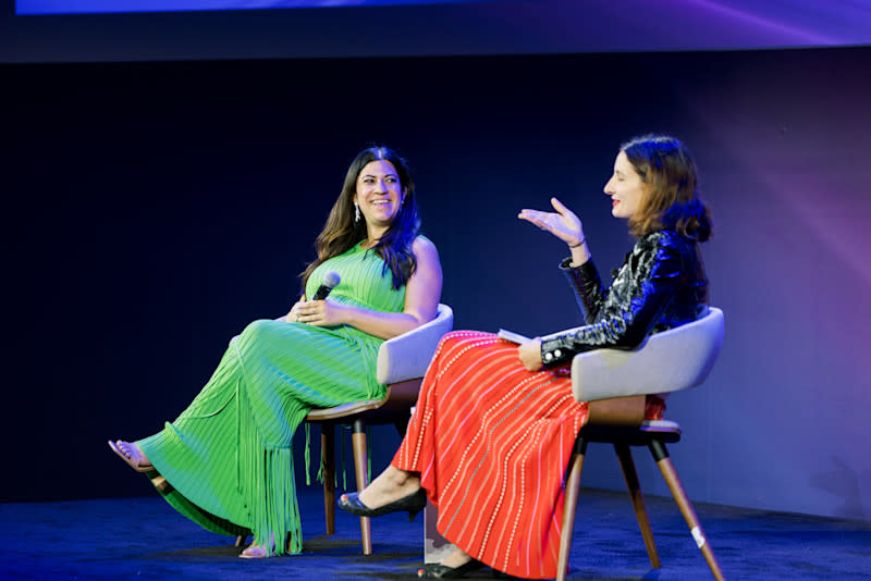 Two women are seated in chairs on a stage, facing each other in conversation. The woman on the left, wearing a long, pleated, bright green dress, is smiling and holding a microphone. The woman on the right, in a red striped skirt and a shiny black jacket, is gesturing with her hand while speaking.