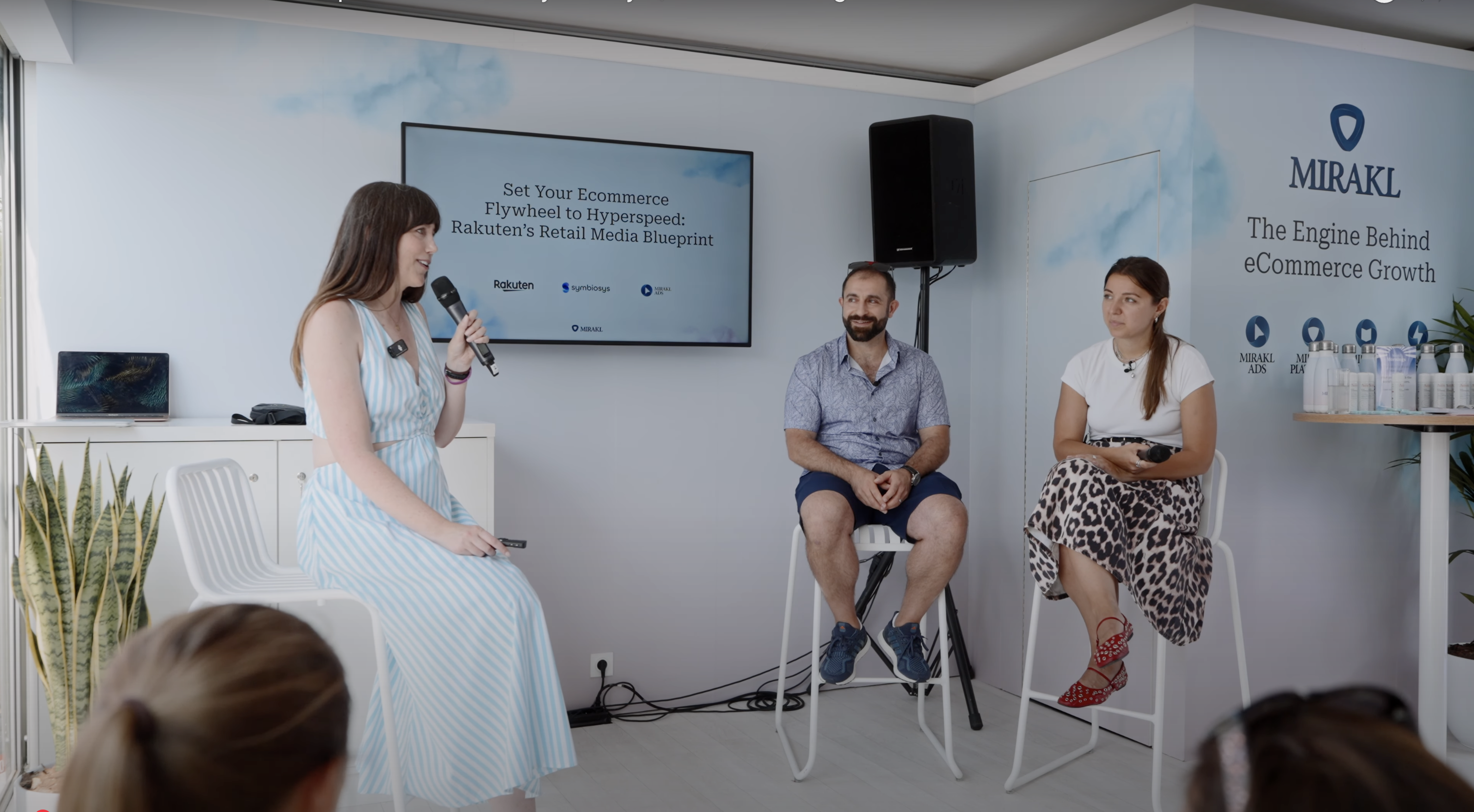 Three people are seated on high stools on a small stage during a Mirakl Mornings session at Cannes. A woman on the left, Anne Hallock, VP, Sales for Mirakl Ads, is speaking into a microphone. In the center, a man with a beard, Bashar Kachachi, Founder and CEO of Symbiosys, is seated. On the right, a woman, Kristina Raptovaia, Rakuten France's Head of Partnerships and Advertising, is seated. The large screen behind them displays the title "Set Your eCommerce Flywheel to Hyperspeed: Rakuten's Retail Media Blueprint," and the event discussed how their collaboration fuels a more connected, performant retail media ecosystem. The wall behind the stage features a cloud pattern and the "MIRAKL" logo.