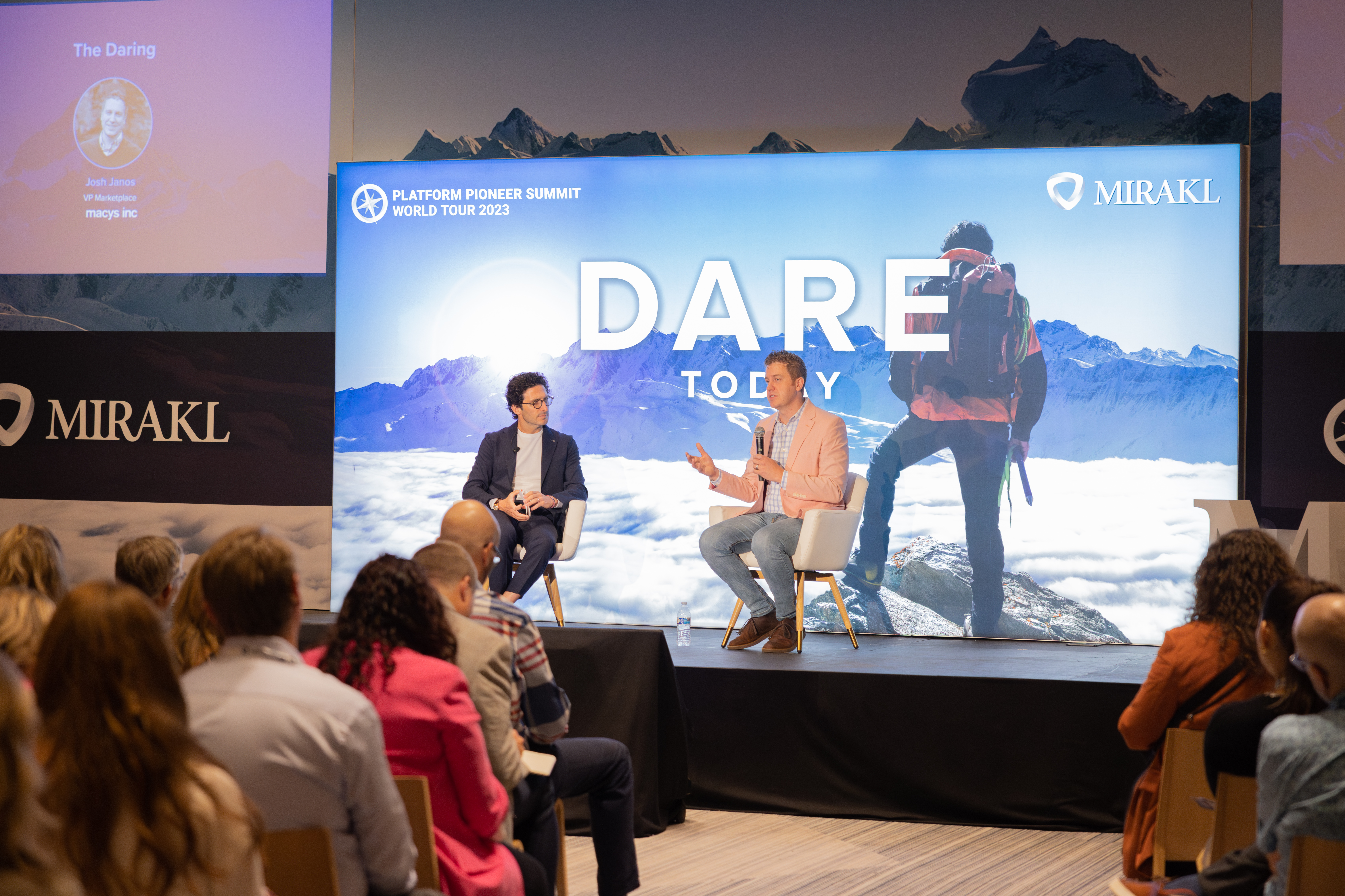 A wide shot of an indoor conference presentation. Two men are seated in chairs on a stage, facing each other and gesturing as they speak. Behind them, a large screen displays "DARE TODAY" and a graphic of a person hiking in mountains, along with "PLATFORM PIONEER SUMMIT WORLD TOUR 2023" and the "MIRAKL" logo. In the foreground, an audience is seated with their backs to the camera, attentively watching the presentation. To the left, a smaller screen shows a speaker's profile with "The Daring" and "MIRAKL" branding.