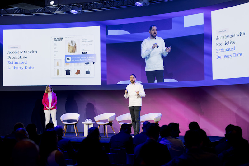 A male presenter in a white sweater and dark pants stands on a stage, speaking into a microphone. Two large screens behind him display the text "Accelerate with Predictive Estimated Delivery Date." The left screen also shows a product page for a jacket on an e-commerce website. A woman in a pink jacket stands to the left of the stage, and several people are seated in the audience.