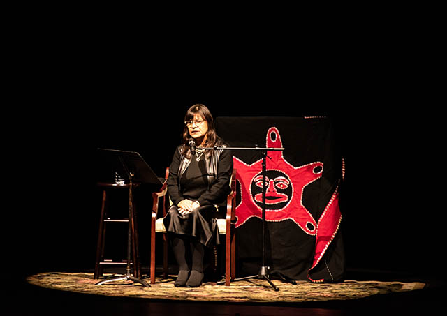 An woman sits on a stage speaking into a microphone, she is wearing black clothing, and is illuminated by a spotlight. There is a red Indigenous art behind her chair. 
