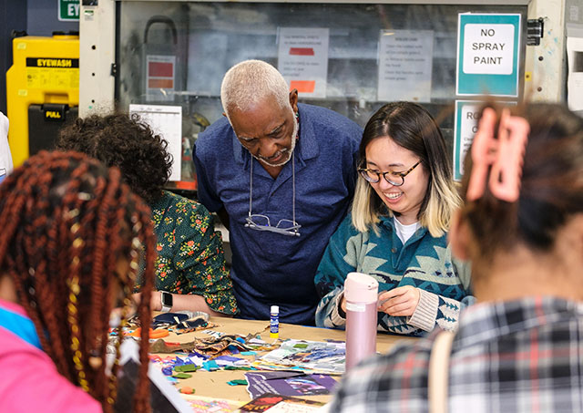 A group of people are gathered around a table covered with various art supplies. They appear to be engaged in a collaborative art project. 