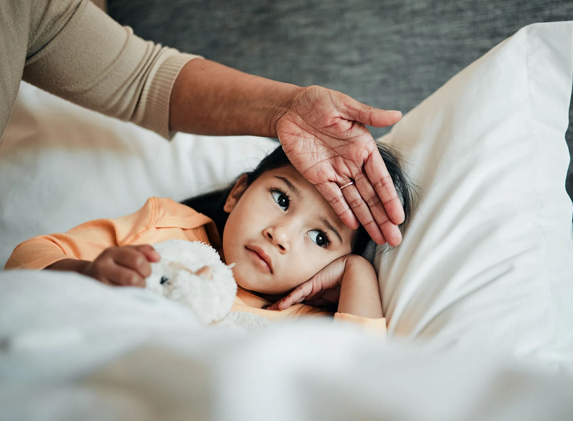 A young child lying in bed with a parent’s hand gently resting on their forehead to check for a fever