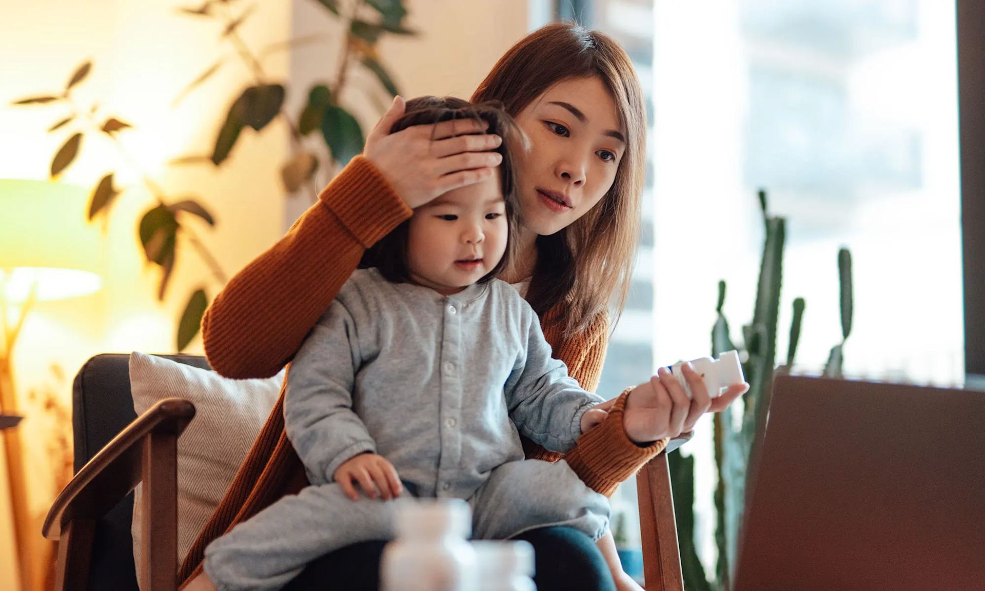 A mother checks her toddler’s forehead while reviewing health information on her laptop to understand their symptoms.