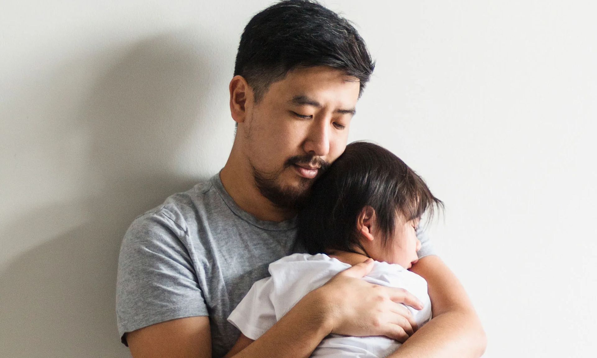 A father comforts and holds a young, sick child in his arms. The child appears to be sleeping, resting his head on his father’s shoulder.