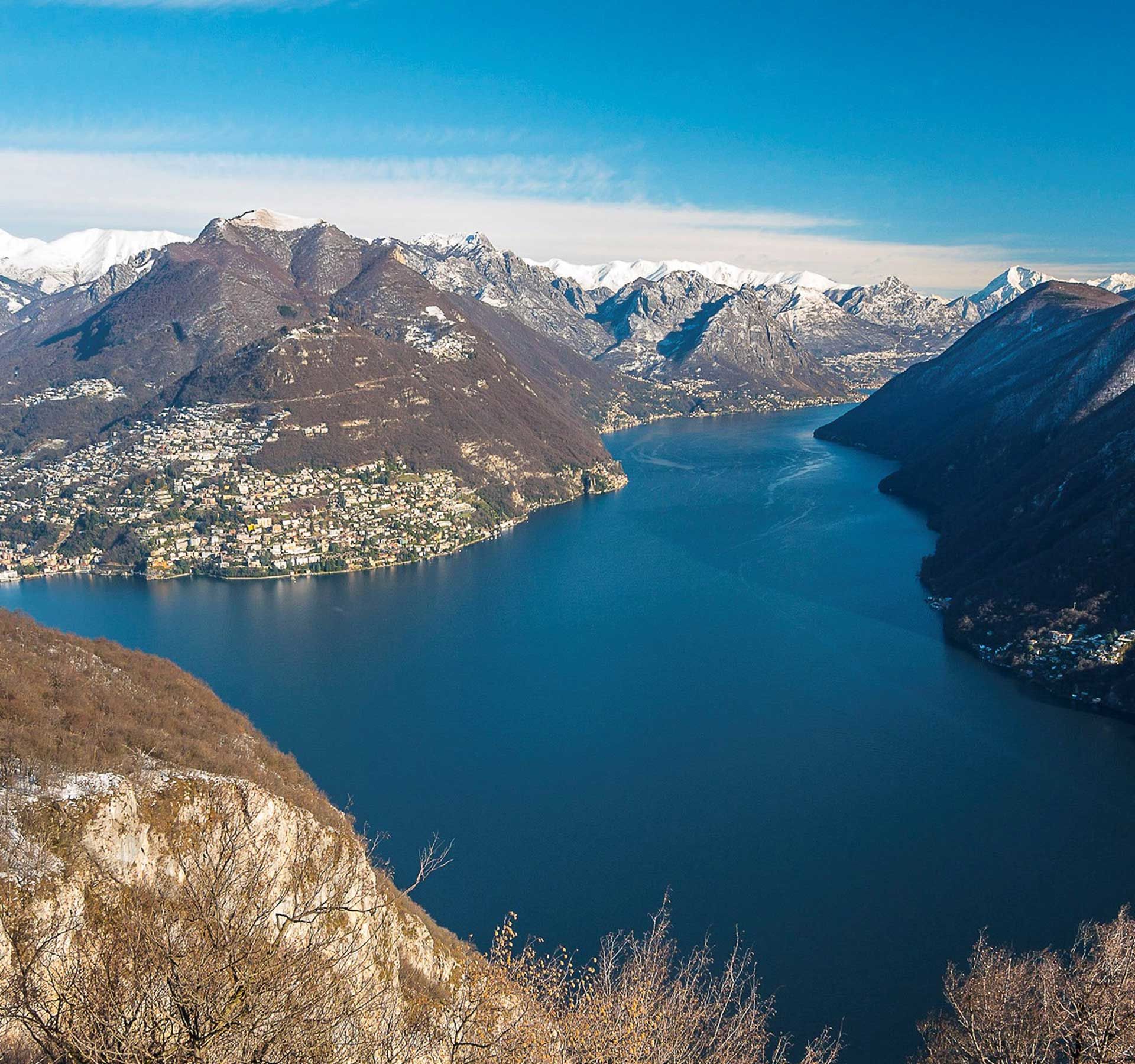 Aerial view of Monte San Salvatore in winter with lightly snow-covered mountains and a view of the deep blue Laggio Maggiore.