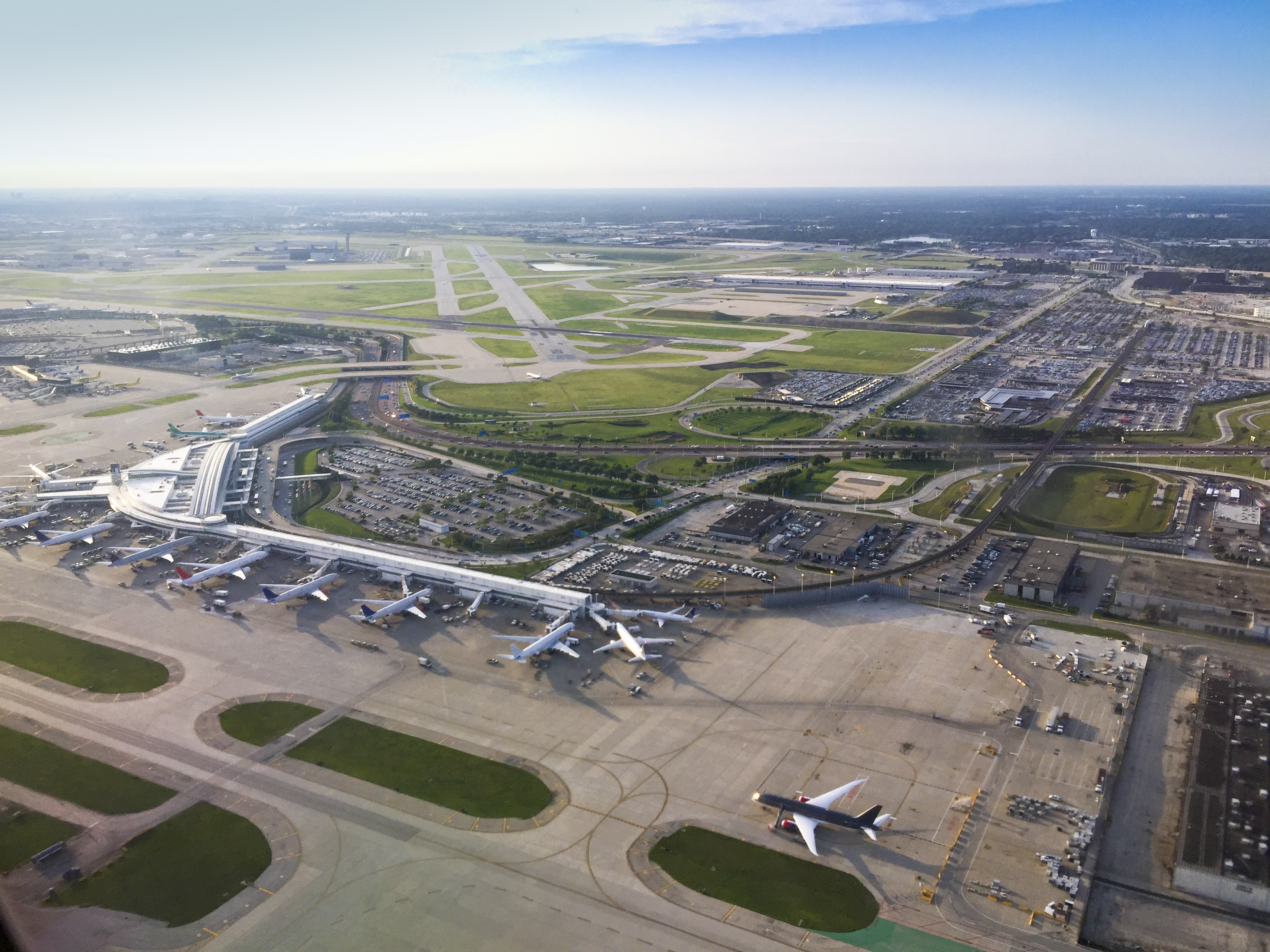 Aerial view of a busy airport terminal with multiple planes parked at gates, runways stretching into the distance, and surrounding parking lots and roads.