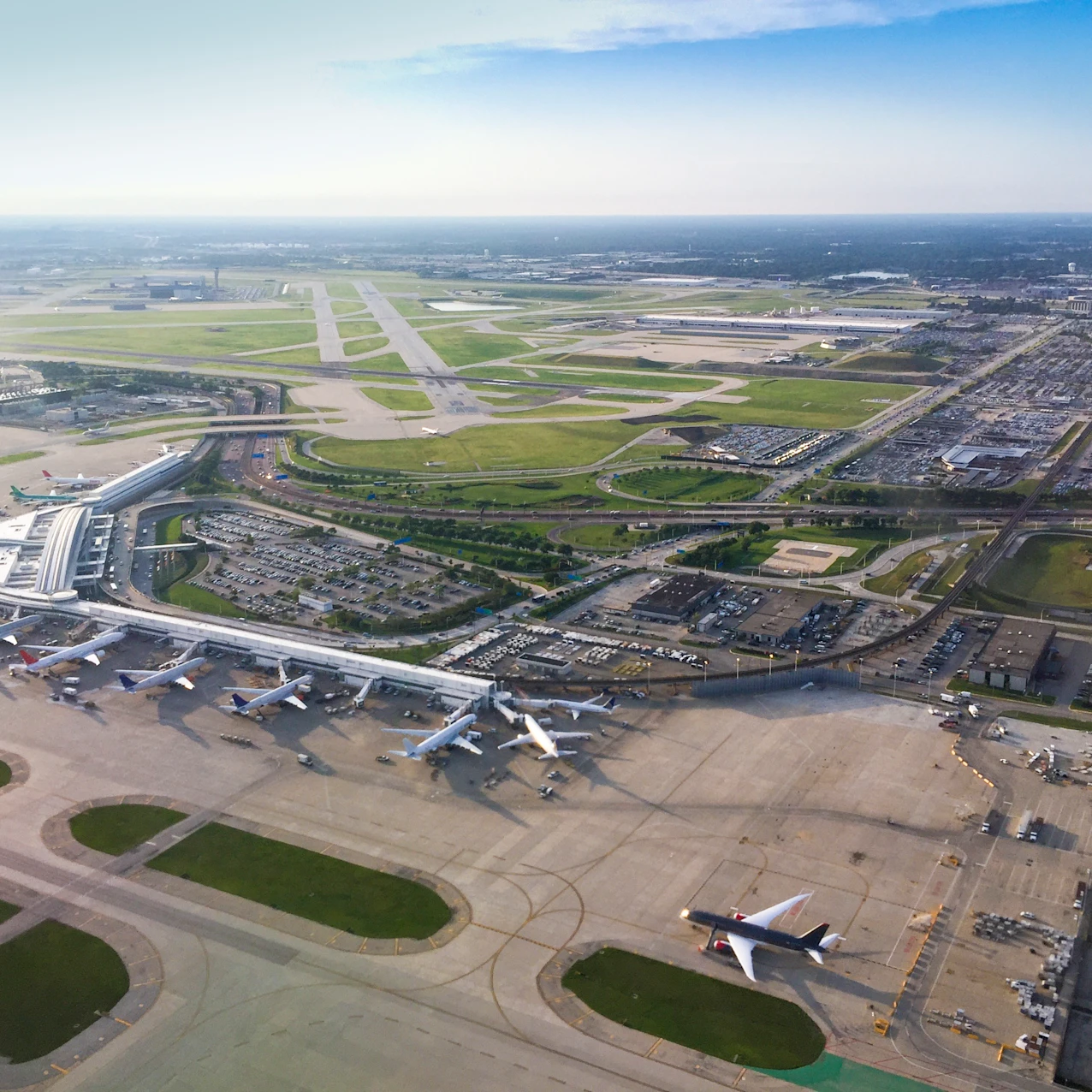 Aerial view of a busy airport terminal with multiple planes parked at gates, runways stretching into the distance, and surrounding parking lots and roads.