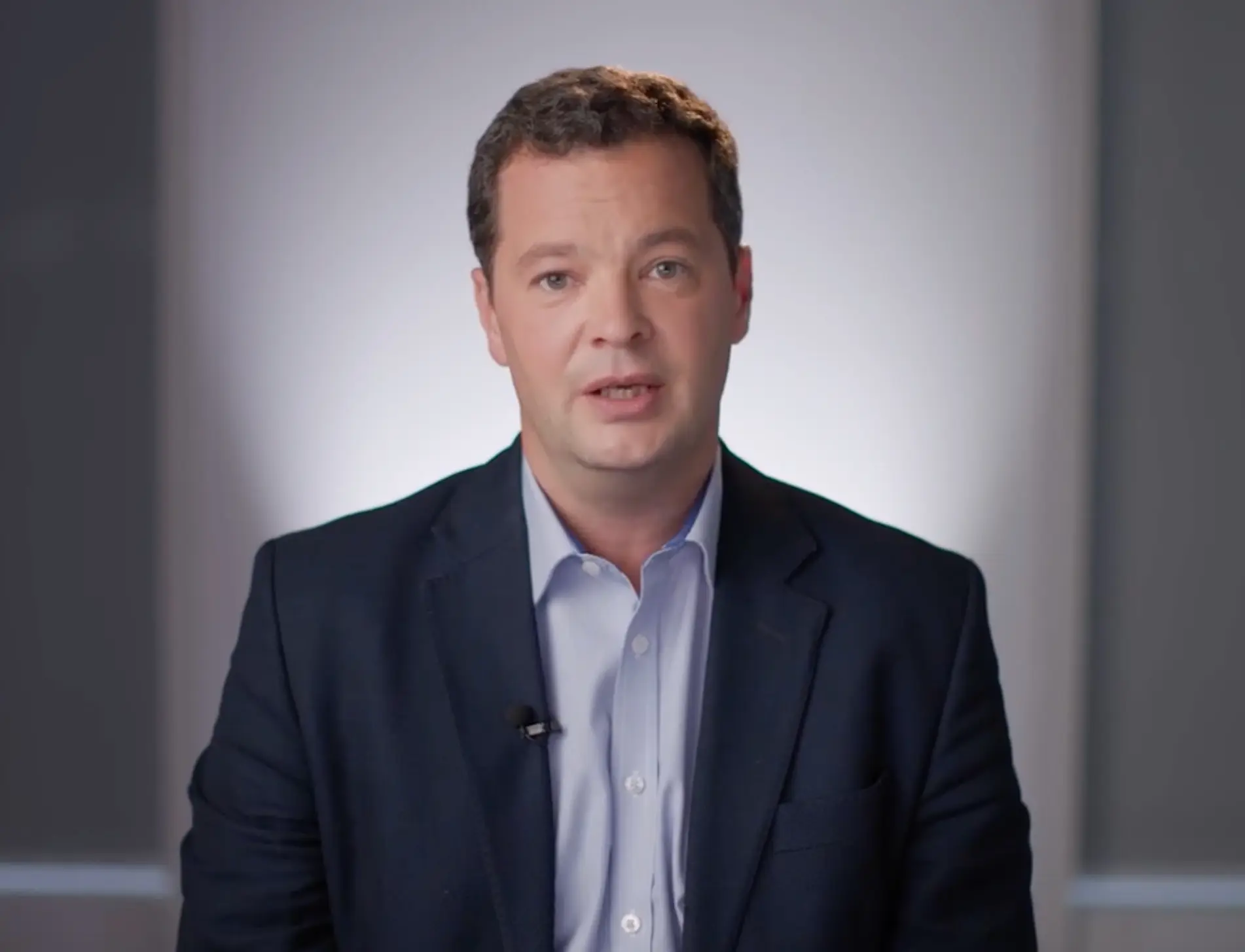 Man wearing a dark blazer and light blue shirt, seated against a plain, softly lit background.