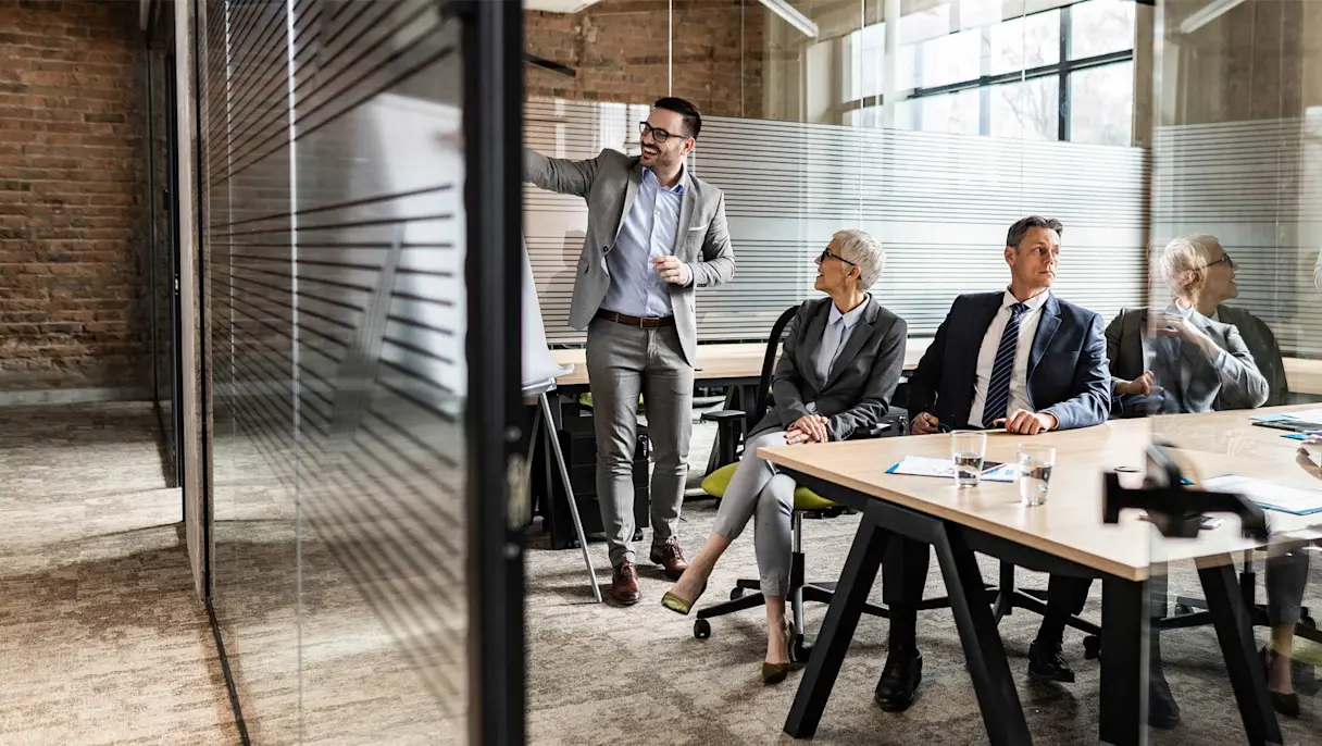 Professional business meeting in a modern office with a presenter leading a discussion while colleagues are seated around a desk.