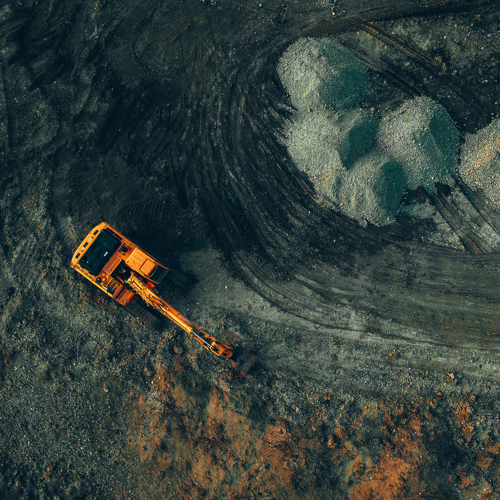 Vue aérienne d’une pelle mécanique orange travaillant sur un chantier avec terre et gravier, entourée de traces de pneus et de tas de pierres.