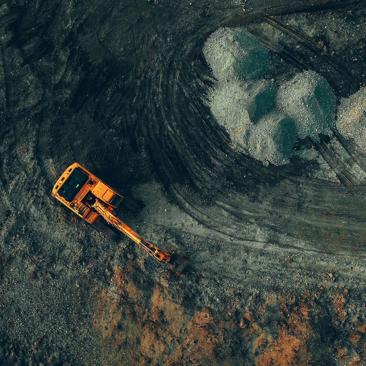 Vue aérienne d’une pelle mécanique orange travaillant sur un chantier avec terre et gravier, entourée de traces de pneus et de tas de pierres.