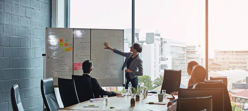 A professional pointing to a whiteboard in a meeting room with other professionals